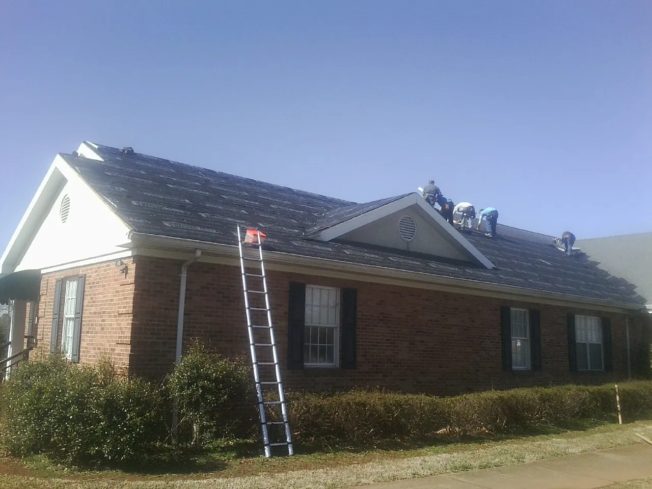 Crew tearing shingles off a brick ranch roof with a ladder set — Metal Roof Repair work by our crew in Adrian
